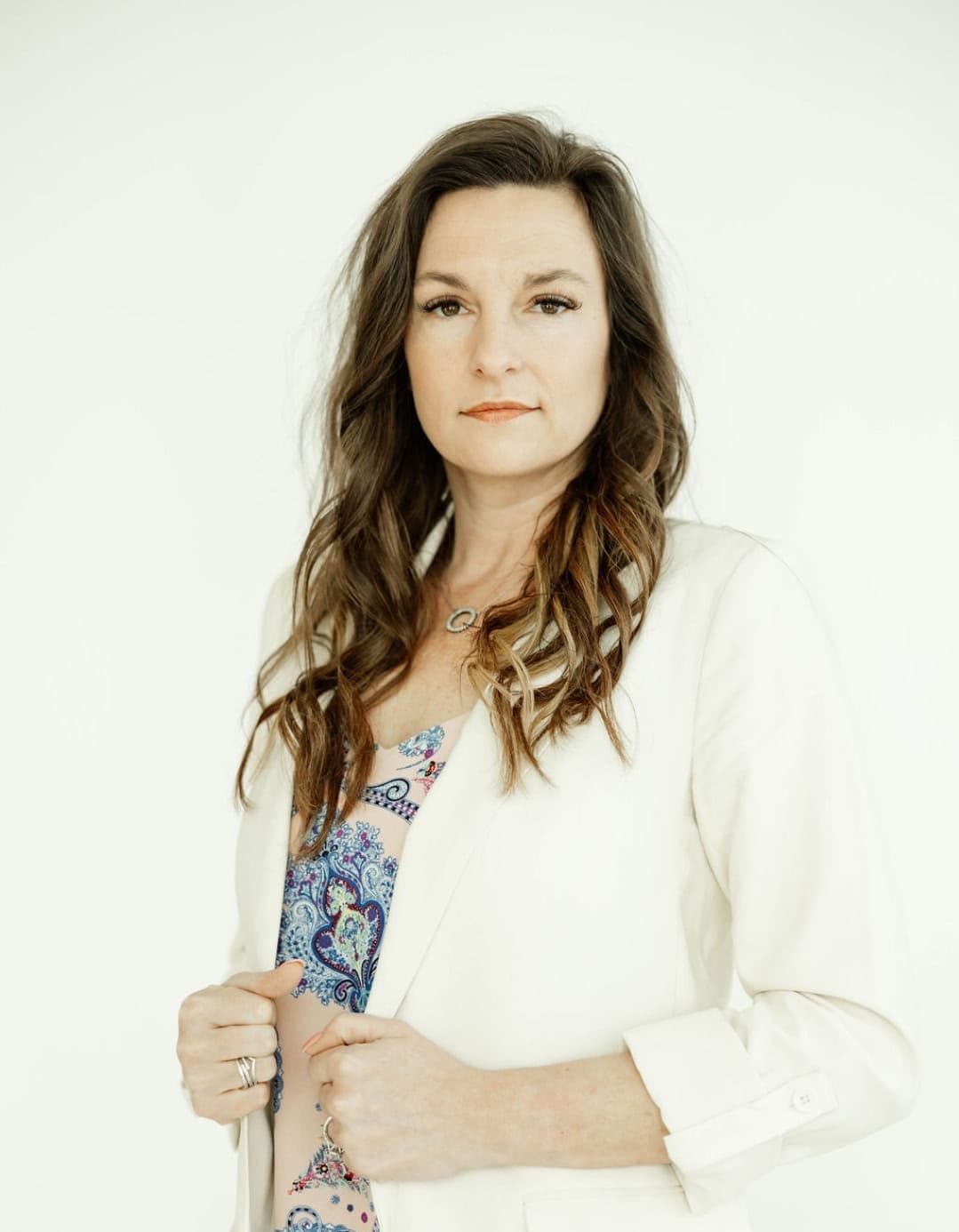 Woman with long wavy hair wearing a white blazer and patterned top against white background.